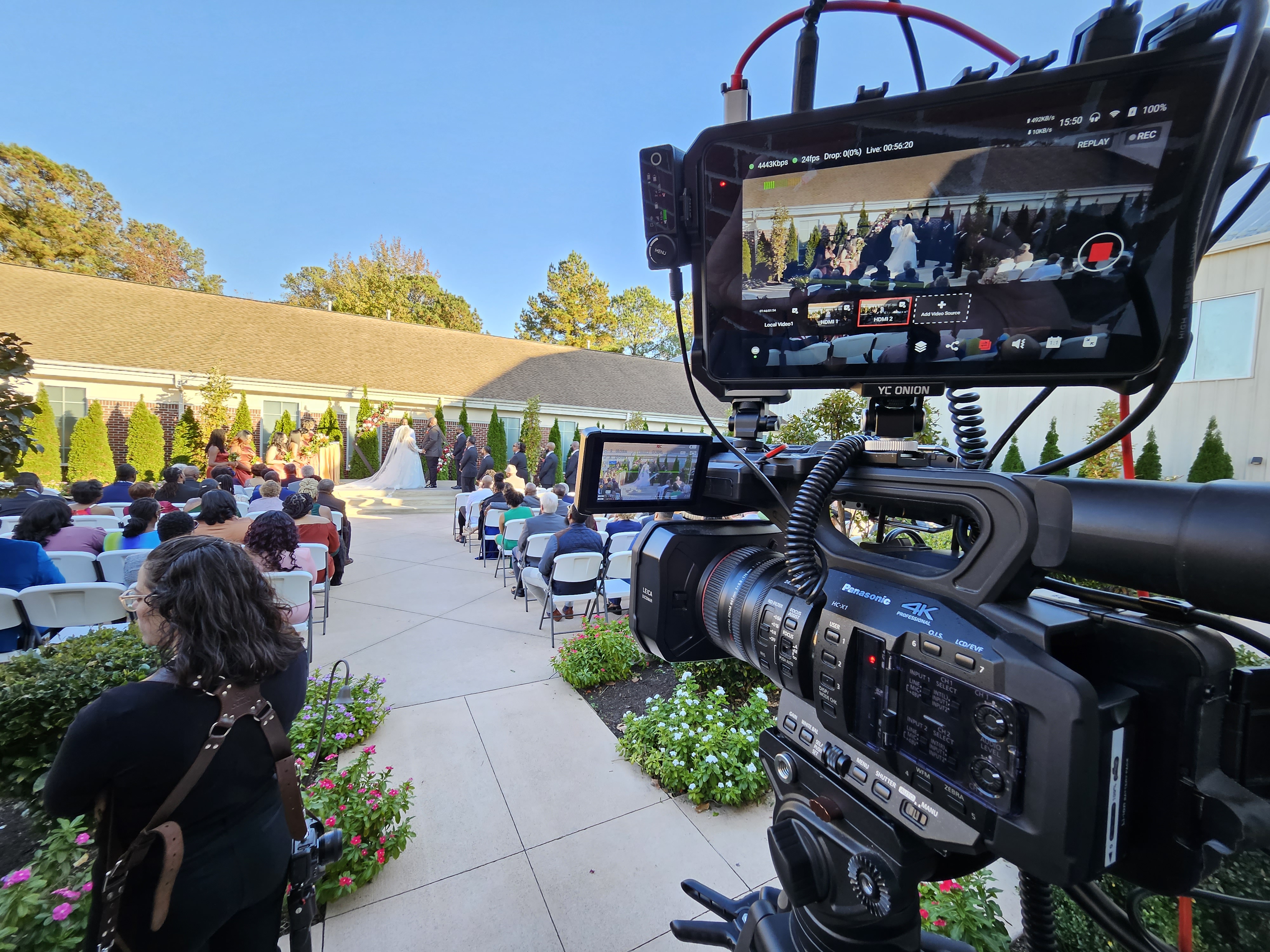 A wedding ceremony taking place outdoors with guests seated in rows, and a video camera focused on the couple at the altar.