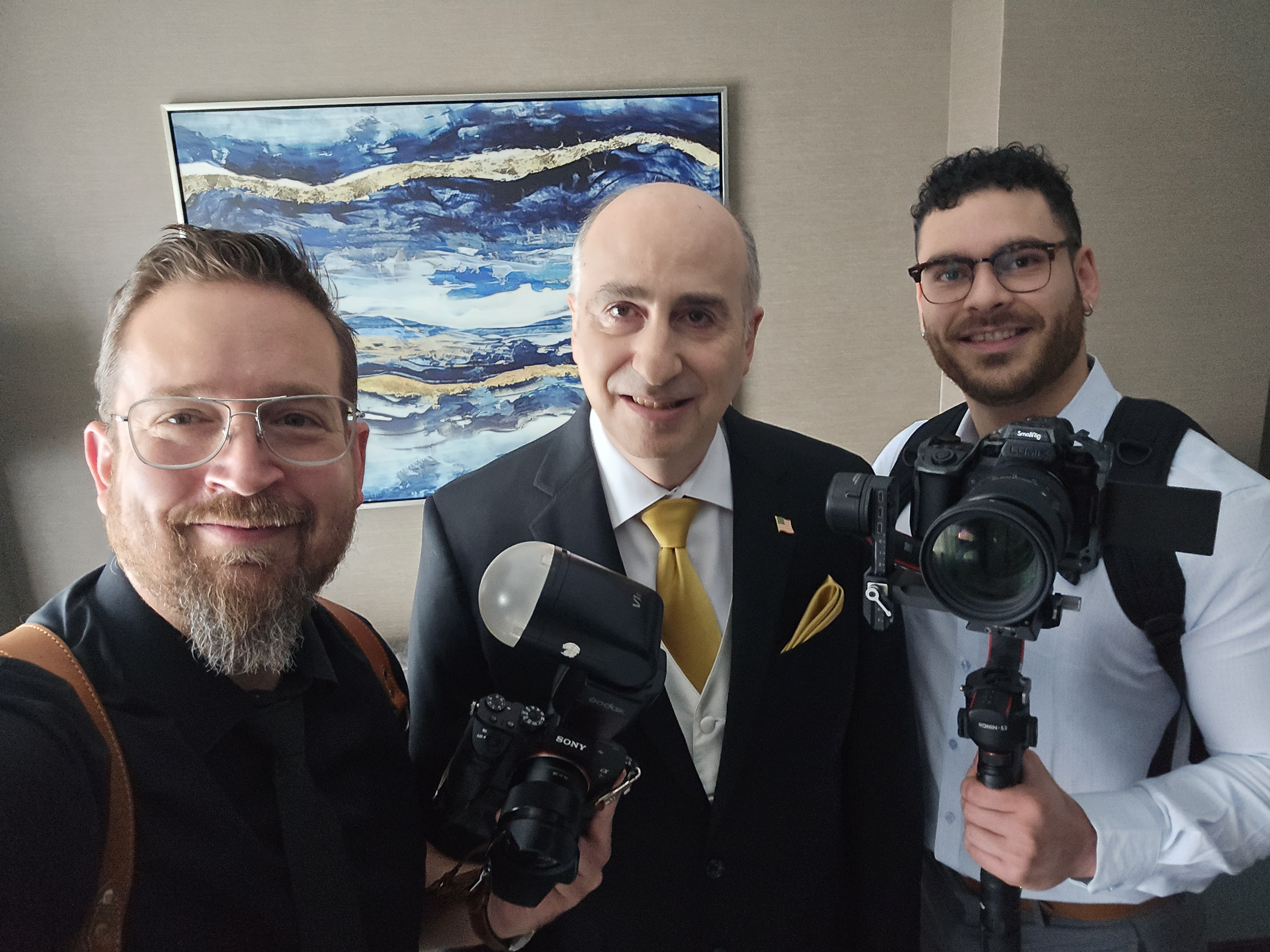 Three men pose for a selfie in a well-lit room, with one wearing a black suit and yellow tie, while others hold cameras and flash equipment.