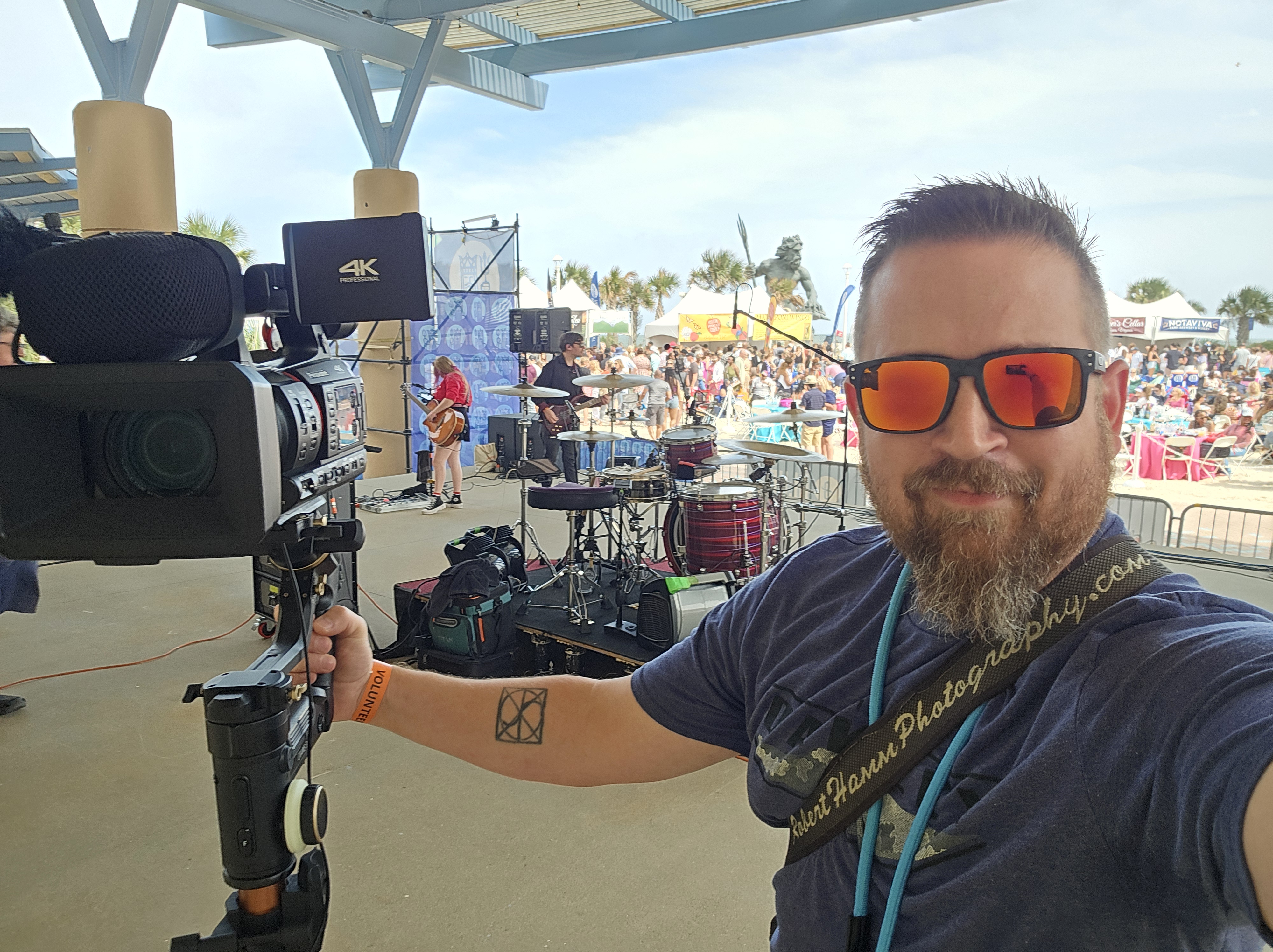 A man with sunglasses holds a camera, smiling for a selfie. In the background, a live music band is performing at an outdoor event, with a large crowd and festival setup visible.