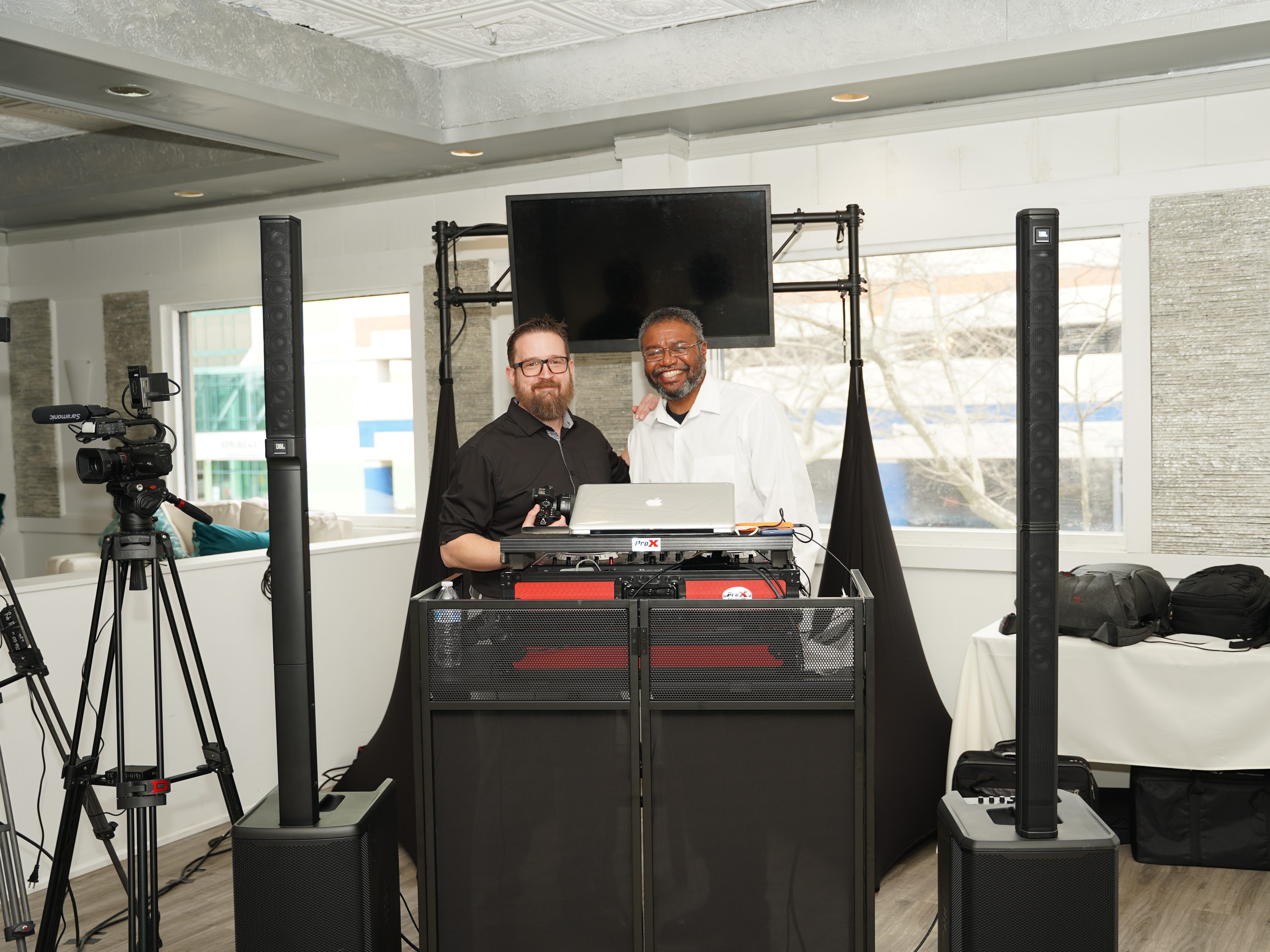 Two men standing behind a DJ setup with speakers and cameras in a well-lit room.