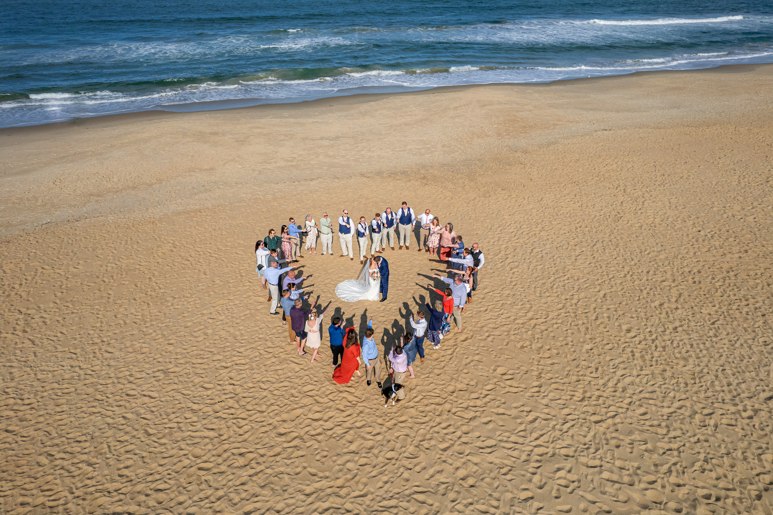 Aerial view of a wedding couple surrounded by friends and family forming a heart shape on a sandy beach.