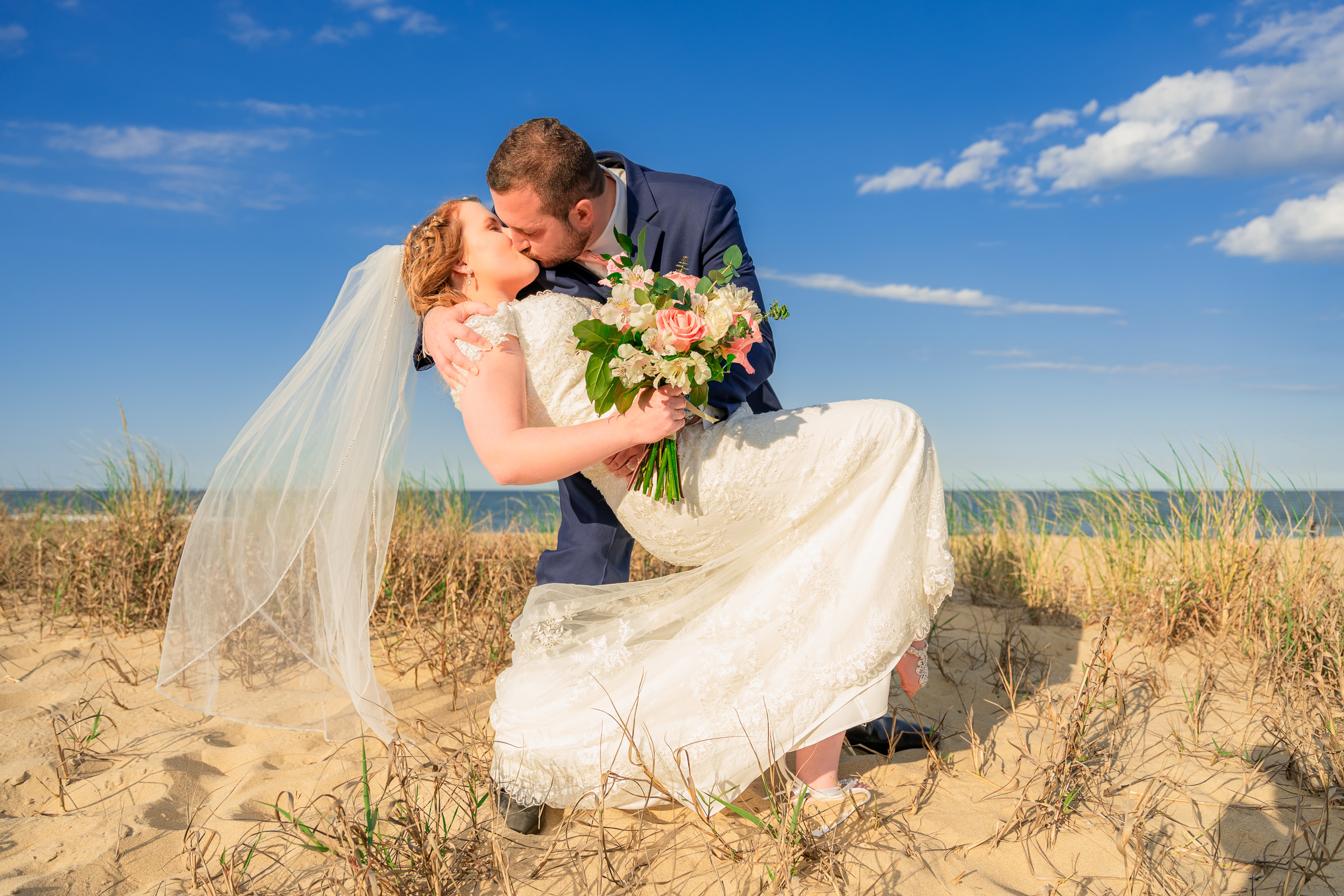 Bride and groom sharing a kiss on the beach, with the bride in a white lace wedding gown and the groom in a navy suit, holding a bouquet of flowers.