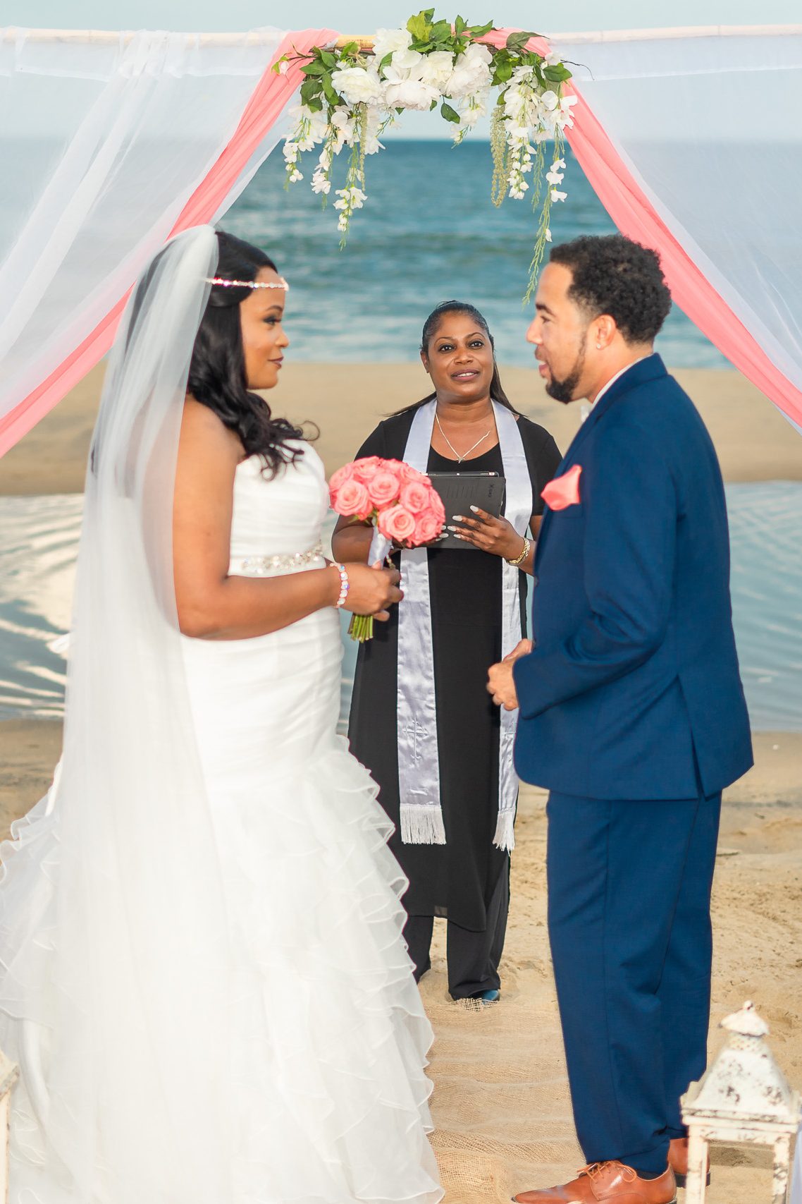 A bride in a white gown holding pink roses stands at an outdoor wedding altar by the beach. A man in a blue suit looks at her while a officiant in a black dress stands between them, overseeing the ceremony.