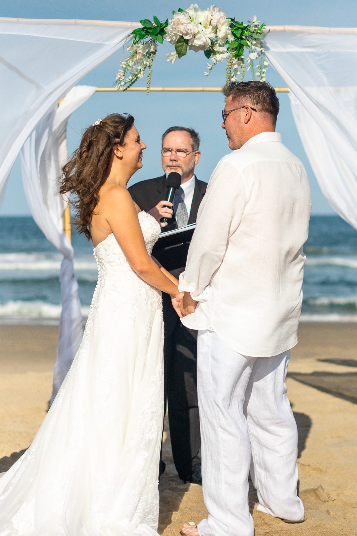 A couple exchanging vows during a beach wedding ceremony, with a floral arch and officiant in the background.