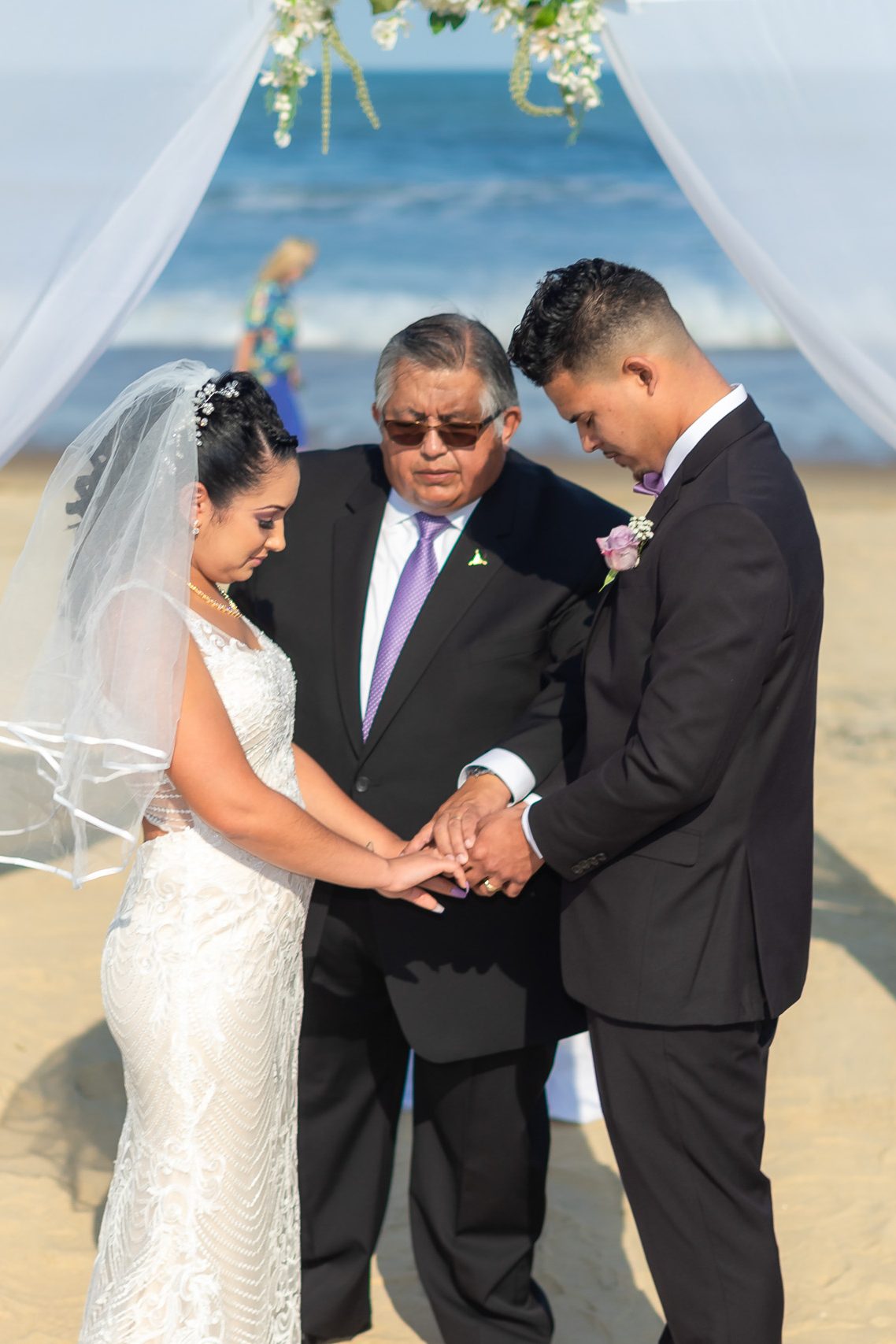 A bride and groom holding hands during their beach wedding ceremony, with an officiant standing between them under a floral arch.