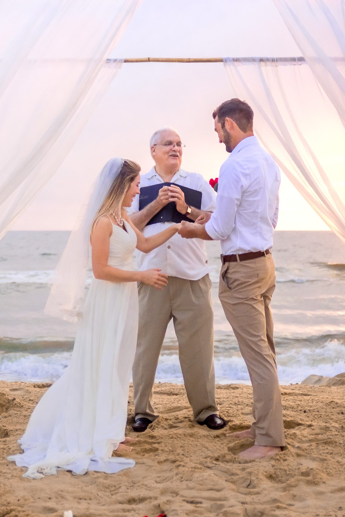 A bride and groom exchanging vows during their beach wedding ceremony, with an officiant standing between them under a decorative canopy.
