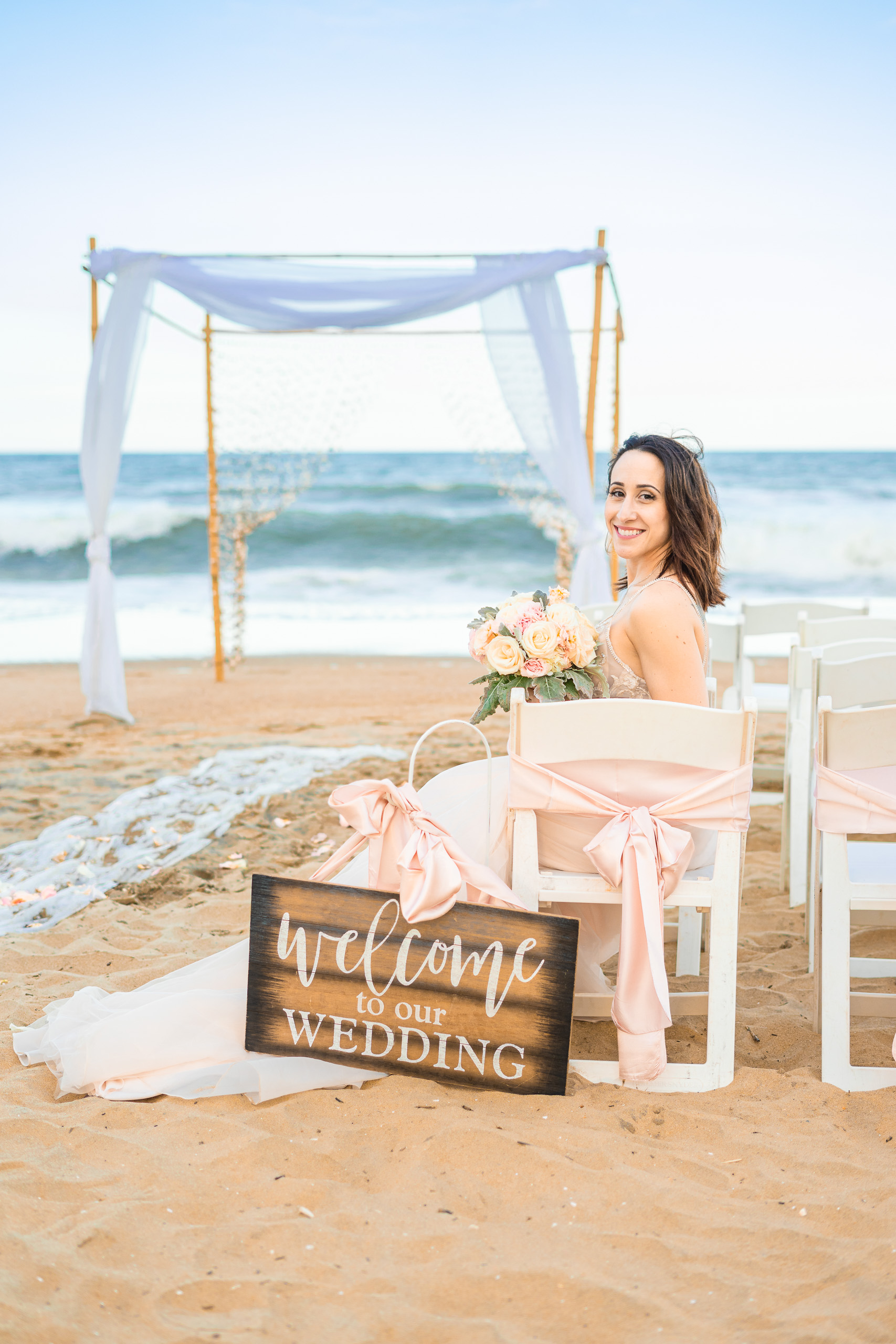 Bride sitting on the beach with a bouquet, smiling at the camera, near a wedding setup with a sign that says 'welcome to our WEDDING'.
