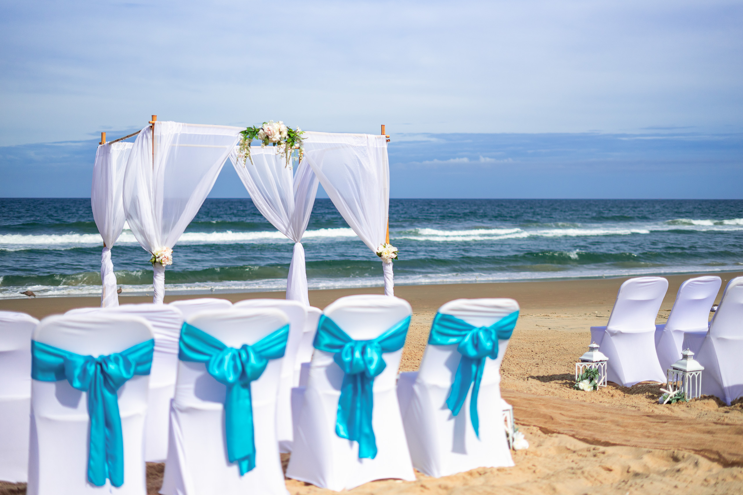 A beach wedding setup featuring white chairs with turquoise bows, facing a floral-adorned canopy by the ocean.