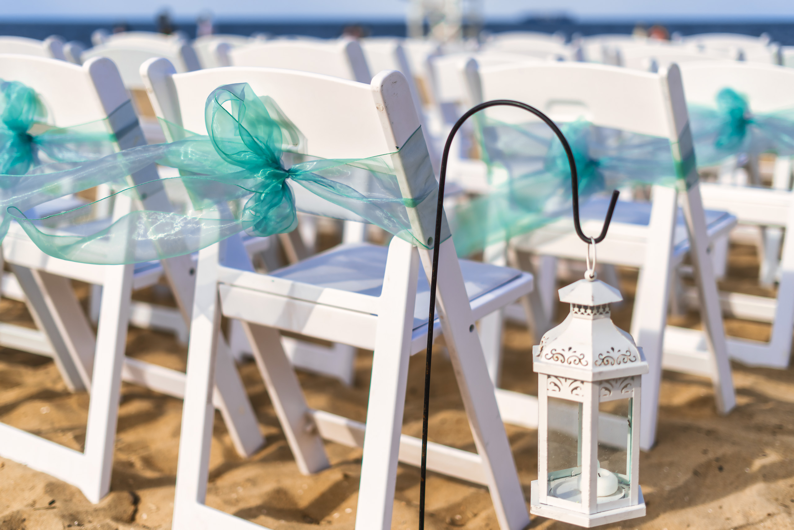 White chairs decorated with aqua ribbons lined up on a sandy beach, with a lantern hanging from one chair.