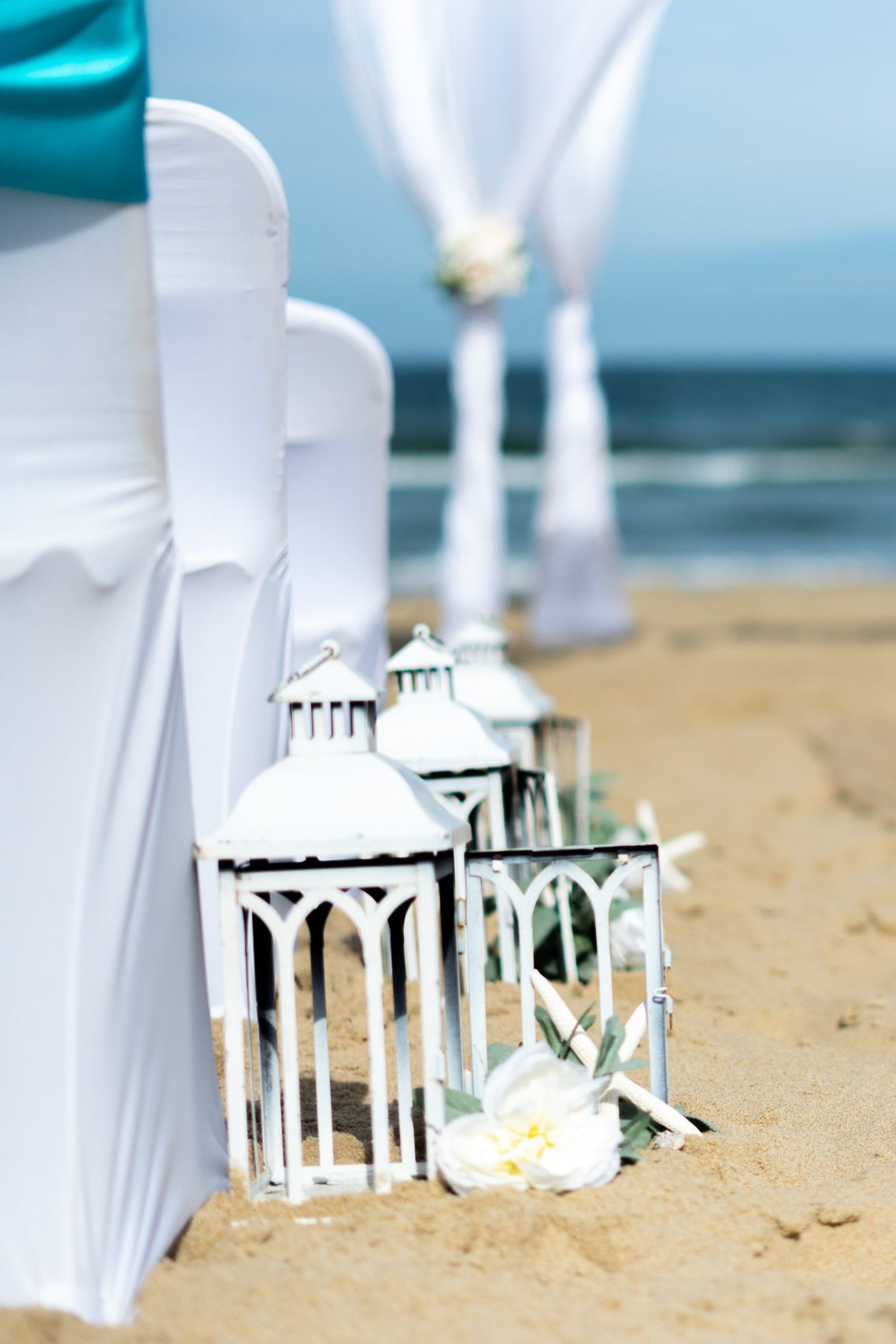 White lanterns and beach chairs set up for a wedding ceremony by the ocean, with floral decorations on the sand.