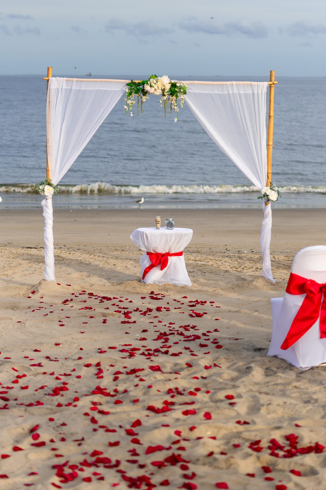 A beach wedding setup featuring a white draped arbor adorned with flowers, a small table with drinks, and scattered rose petals on the sand.