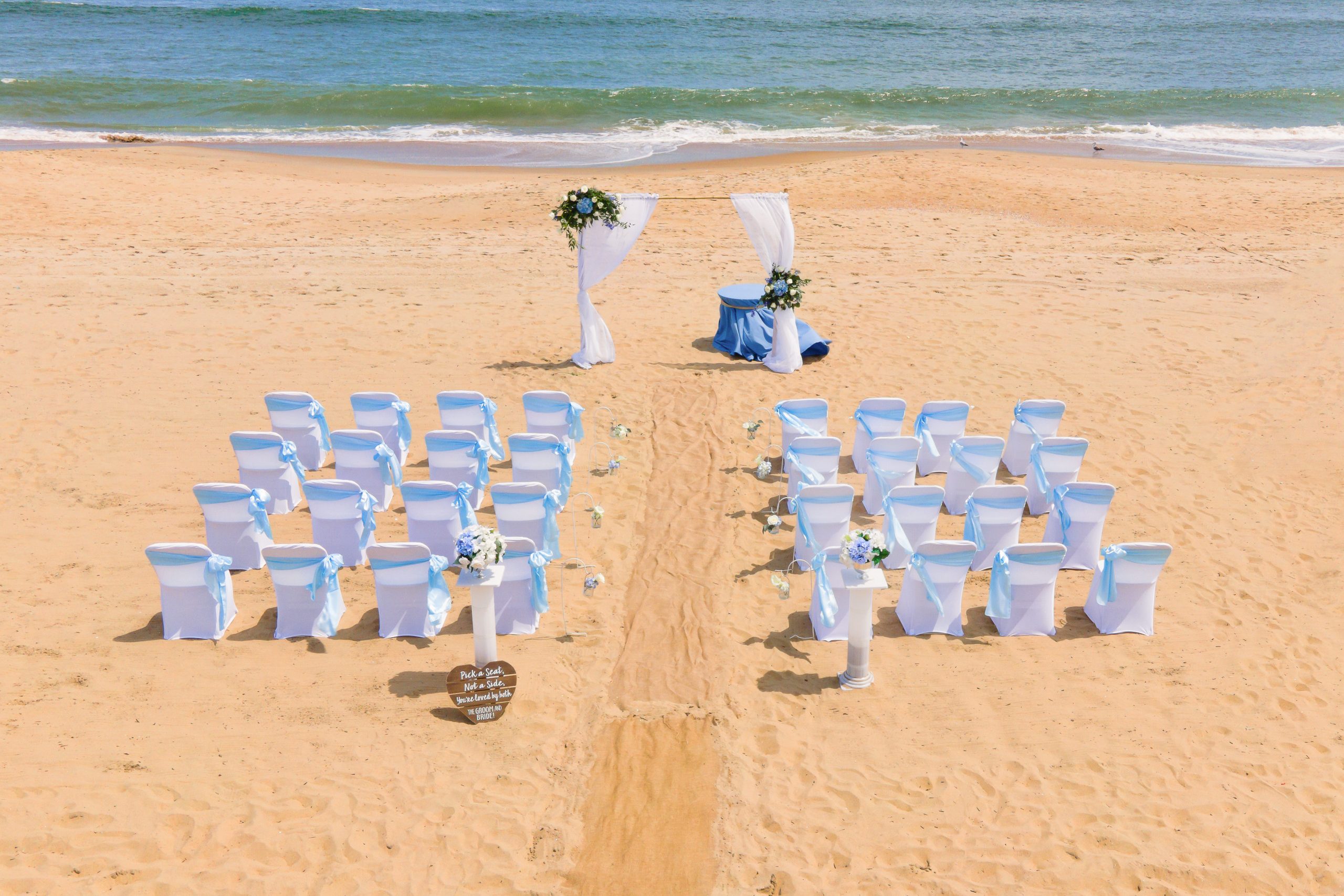 A beach wedding setup with rows of white chairs adorned with blue ribbons, a decorated altar with floral arrangements, and a sandy path leading to it.