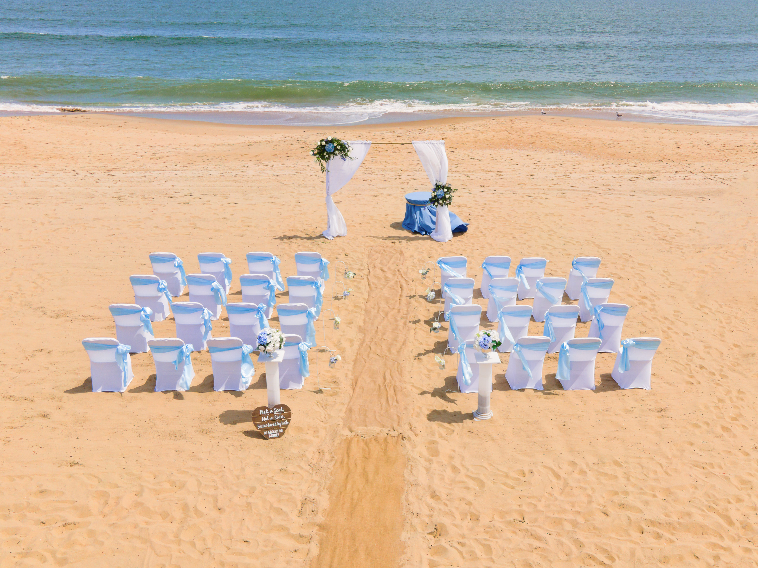 A beach wedding setup featuring rows of elegantly decorated chairs with blue sashes, a floral arch, and a table with decorations, all set against a backdrop of the ocean.