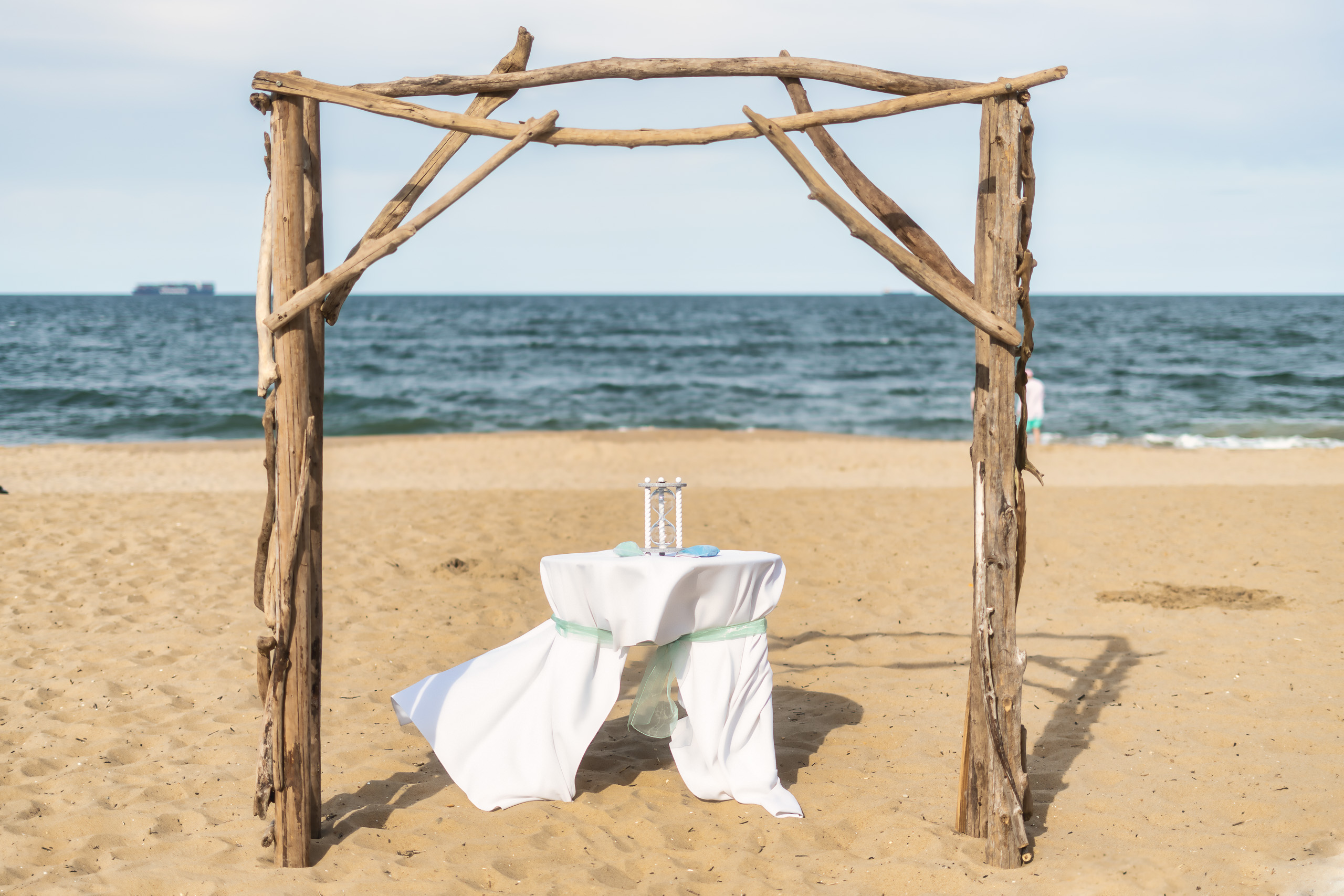 A rustic wooden arch with driftwood frames a small table covered in a white tablecloth at the beach, with the ocean visible in the background.