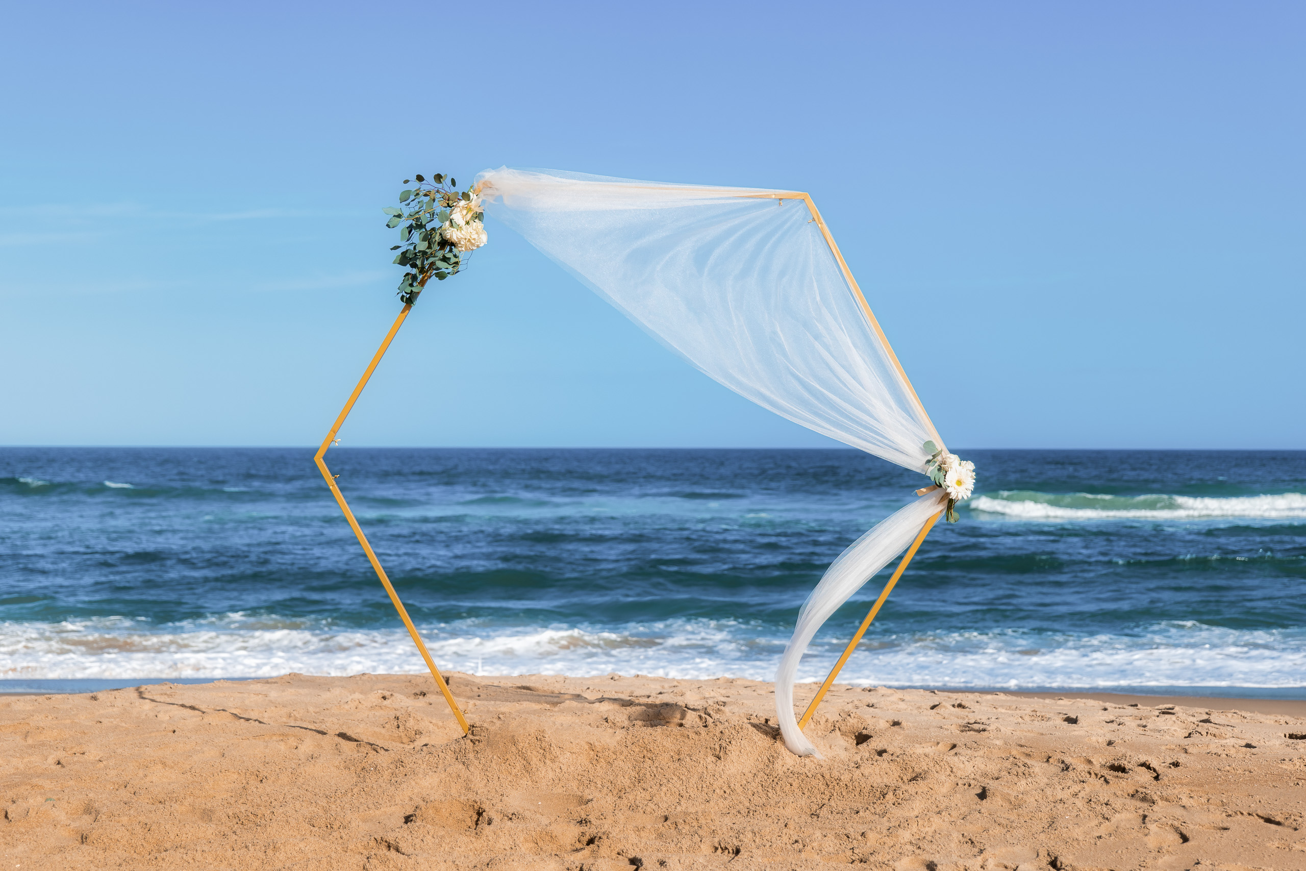 A beach wedding arch decorated with flowers and a flowing white veil, overlooking the ocean under a clear blue sky.