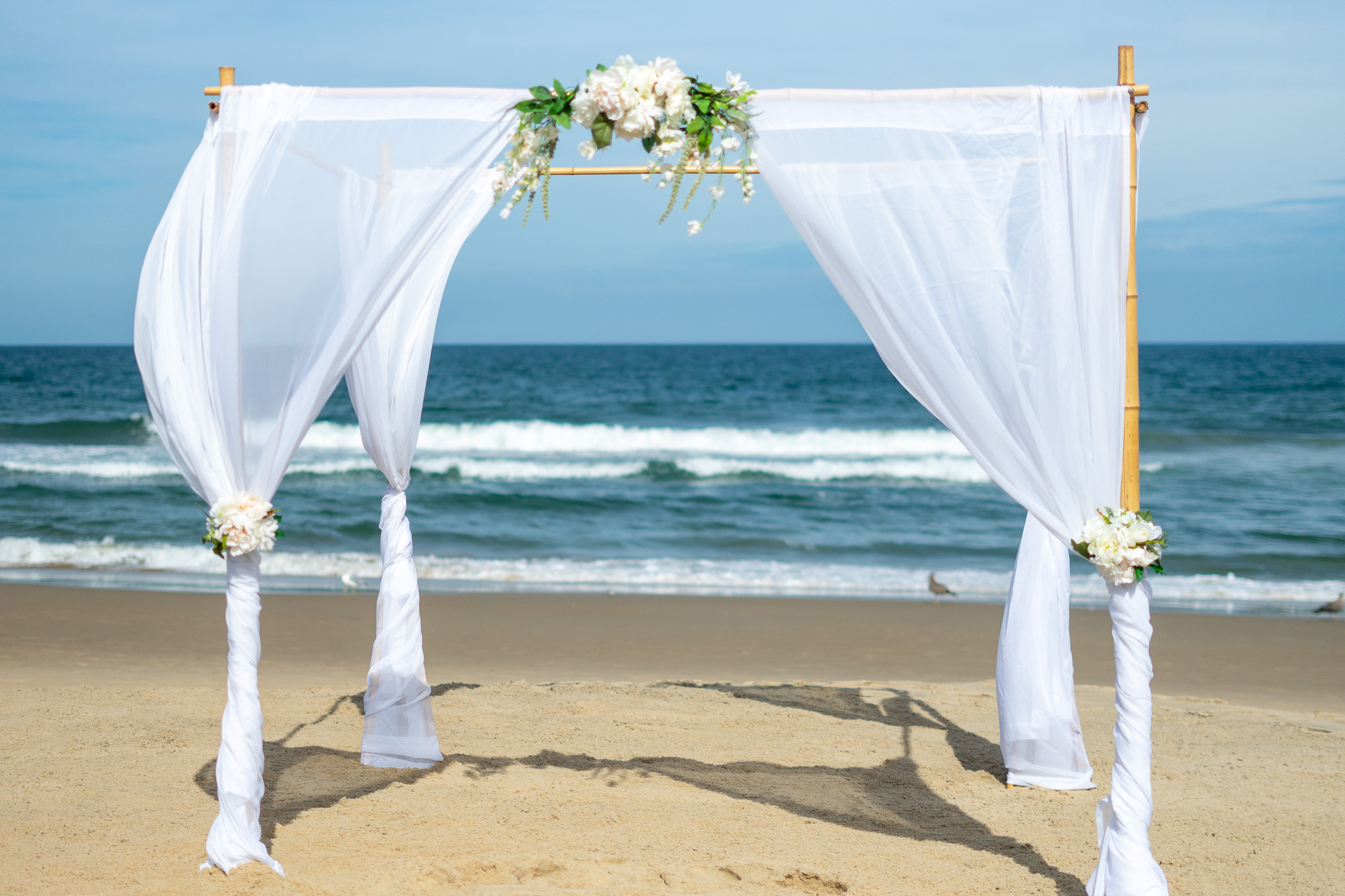 A wedding arch made of bamboo and draped white fabric, adorned with flowers, set against a background of a sandy beach and ocean waves.