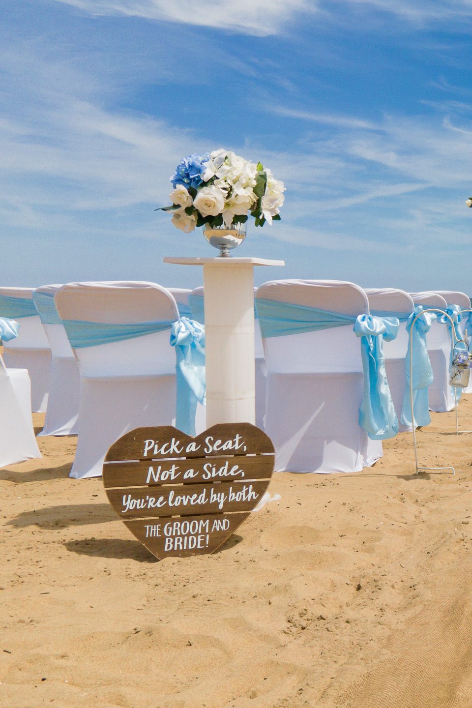 A beach wedding setup featuring white chairs with blue sashes, a decorative floral arrangement on a table, and a heart-shaped sign that reads 'Pick a Seat, Not a Side, You're loved by both the Groom and Bride!'