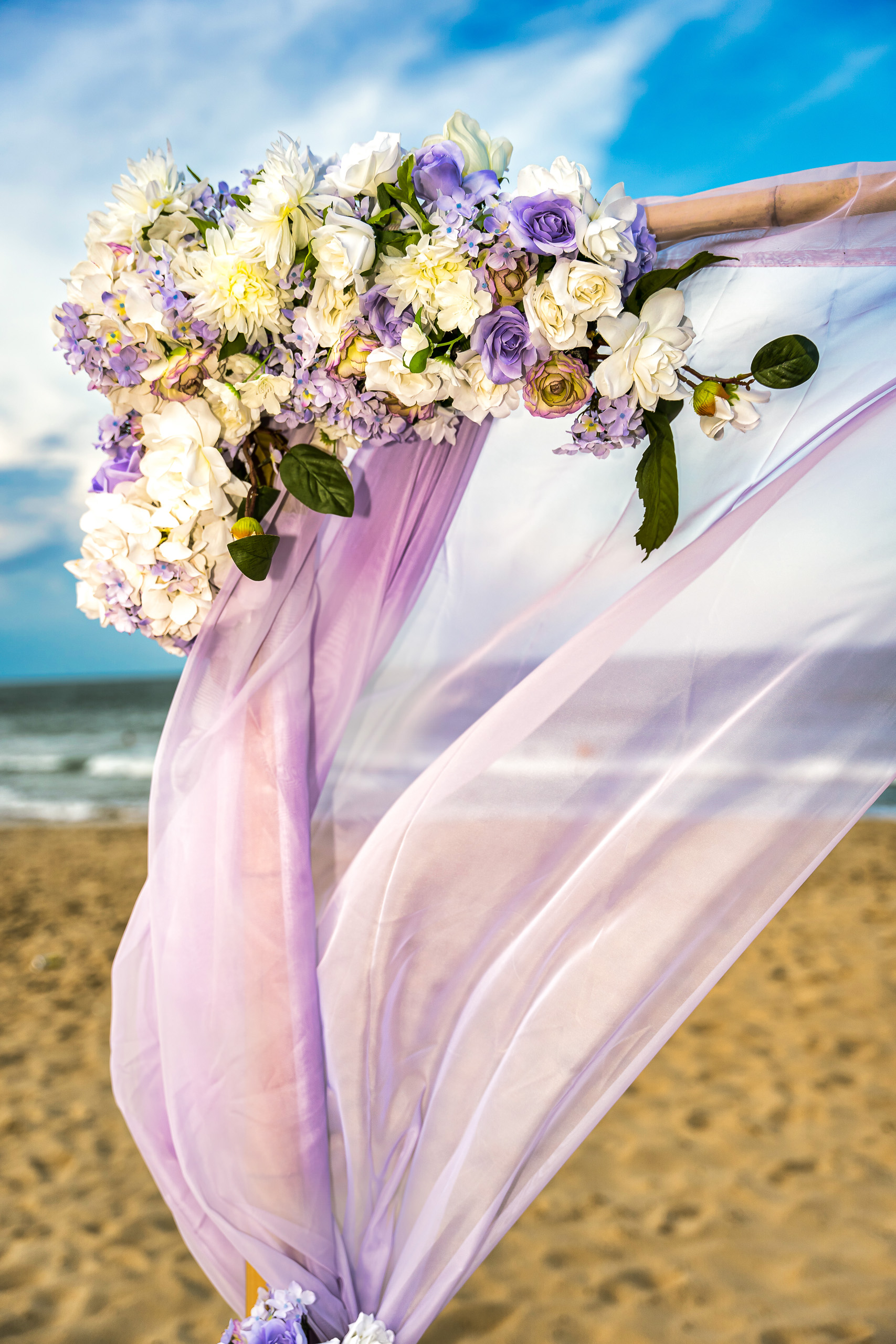 A floral arrangement featuring white, purple, and lavender flowers at the top of a flowing lavender fabric drape, set against a beach background with a cloudy sky.