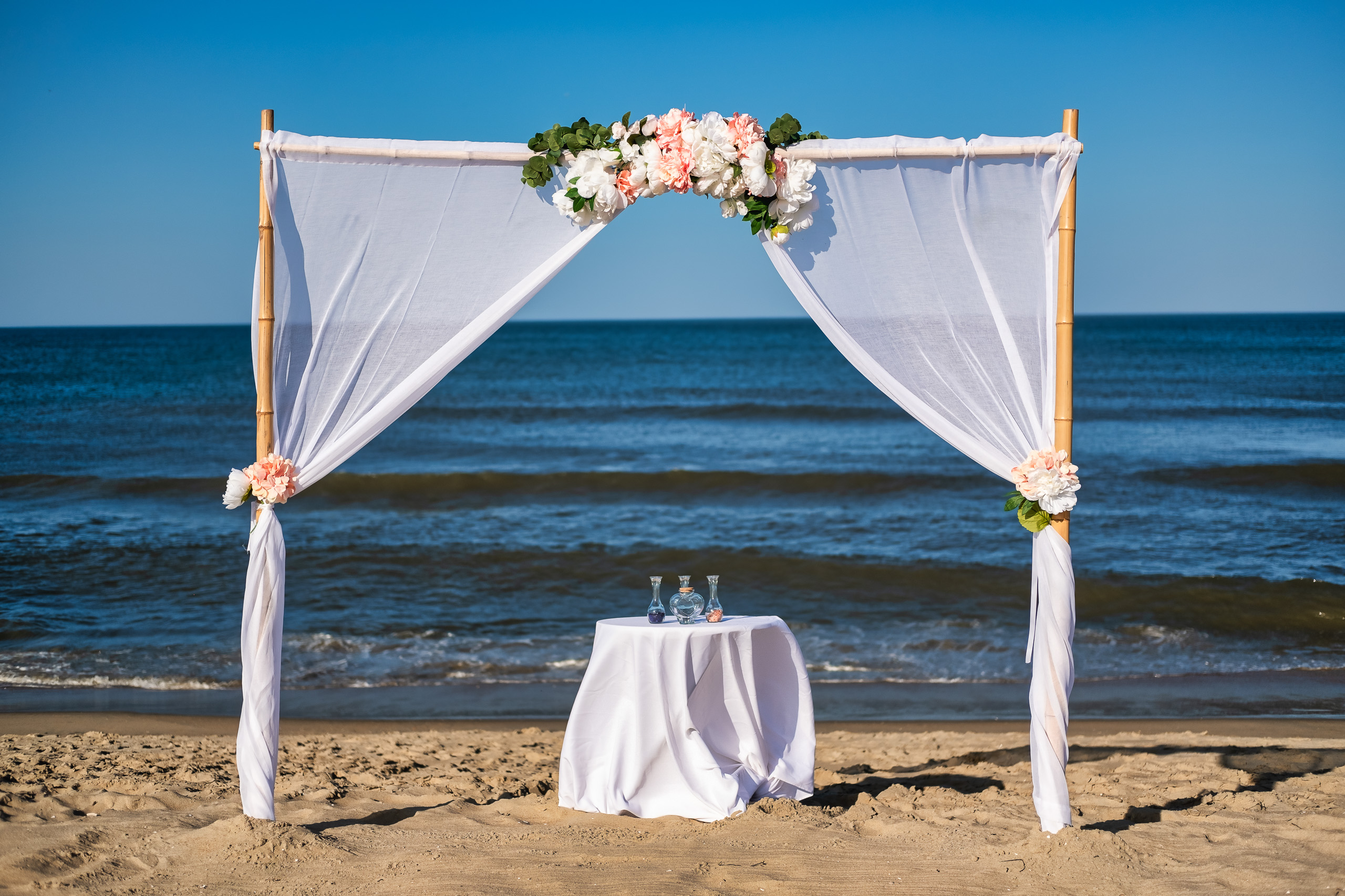 A beachfront wedding altar with white fabric draped on bamboo poles, adorned with flowers, and a small table with glass containers beside it.